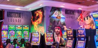 Section of Aristocrat Gaming Lounge with tiled ceiling, carpeted floor, and a variety of colorful slot machines. Walls feature images of game characters, including a pirate.