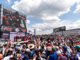 Kyle Bush celebrates with his team in Victory Lane after winning last season's CRC Brakleen 150 NASCAR Craftsman Truck Series Race. A large crowd cheers him on under blue skies.