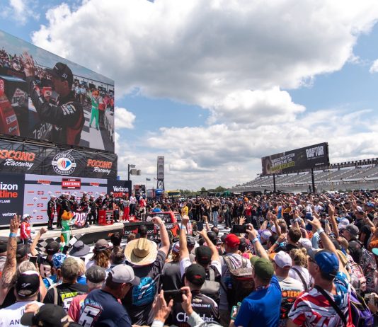 Kyle Bush celebrates with his team in Victory Lane after winning last season's CRC Brakleen 150 NASCAR Craftsman Truck Series Race. A large crowd cheers him on under blue skies.