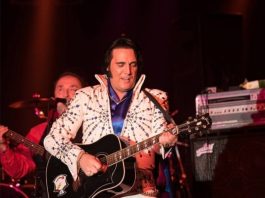 Shawn Klush impersonating Elvis Presley in a white suit and holding a black hollow bodied guitar.